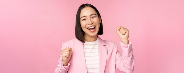 熱狂的なセールスウーマンアジアの企業の女性は、はい、目標を達成し、喜びと笑顔でピンクの背景の上に立って勝利を祝うと言います