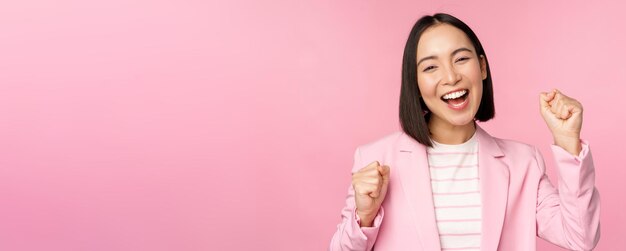열정적인 판매원 아시아 기업 여성은 목표를 달성하고 분홍색 배경 위에 서서 기뻐하고 웃는 모습으로 승리를 축하한다고 말합니다.