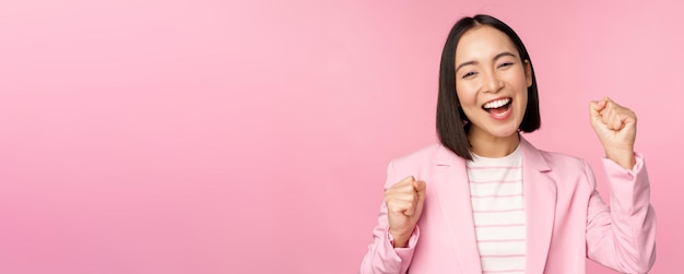 Enthusiastic saleswoman asian corporate woman say yes achieve goal and celebrating triumphing looking with rejoice and smiling standing over pink background