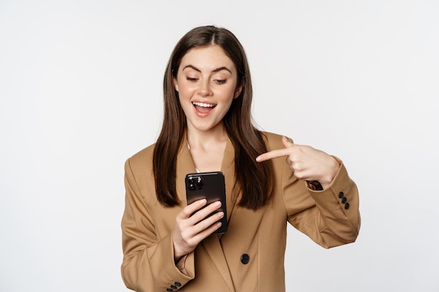Восторженная деловая женщина-продавщица показывает пальцем на мобильный телефон и улыбается, показывая на мобильн...
