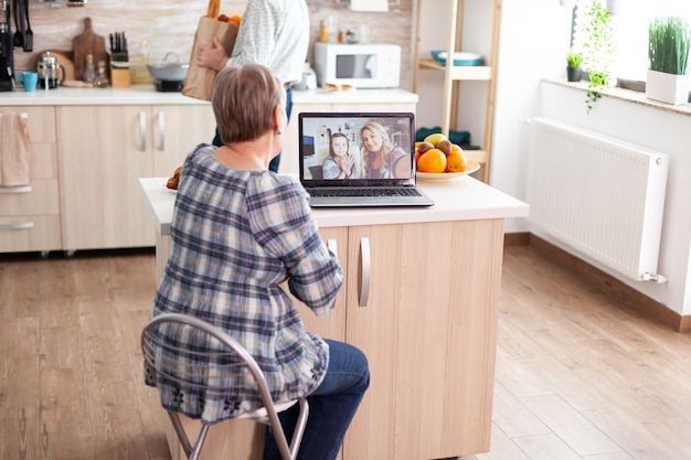 Enthusiastic senior woman talking with family online using laptop webcam during a video conference sitting in kitchen. Videocall with daughter and niece, grandma using modern internet techonolgy.