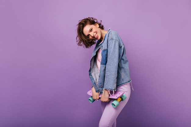 Enthusiastic shorthaired woman in denim jacket holding longboard Winsome white girl with trendy haircut posing with skateboard