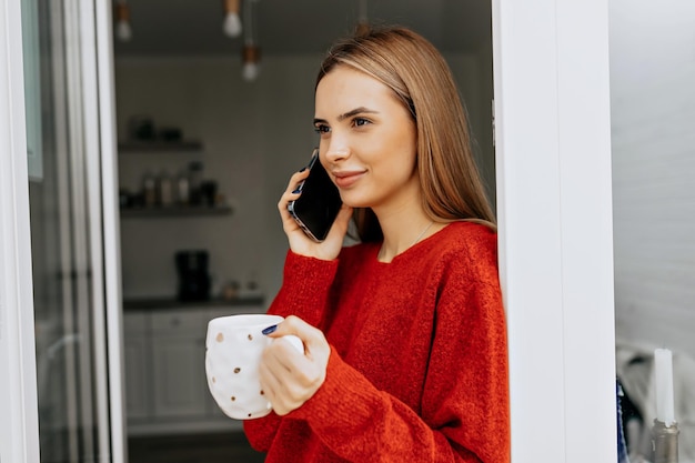 Enthusiastic smiling girl with posing at home with cup of coffee Closeup indoor portrait of enchanting lady in sweater is talking on the phone