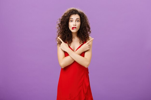 Enthusiastic unsure emotive elegant woman in stylish evening red dress biting lip looking excited pointing in different directions left and right being uncertain and doubtful in making decision.