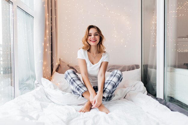 Enthusiastic white girl sitting on bed with legs crossed and expressing positive emotions. Indoor portrait of funny young lady spending time at loggia.