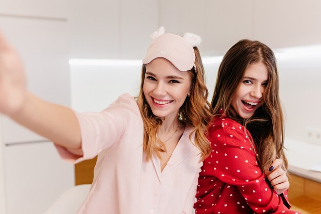 Enthusiastic white girl in trendy night-suit posing in light room with sister. Portrait of laughing curly female model making selfie.