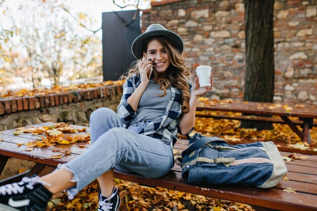 Enthusiastic white lady with long hairstyle sitting on picnic table with blue backpack