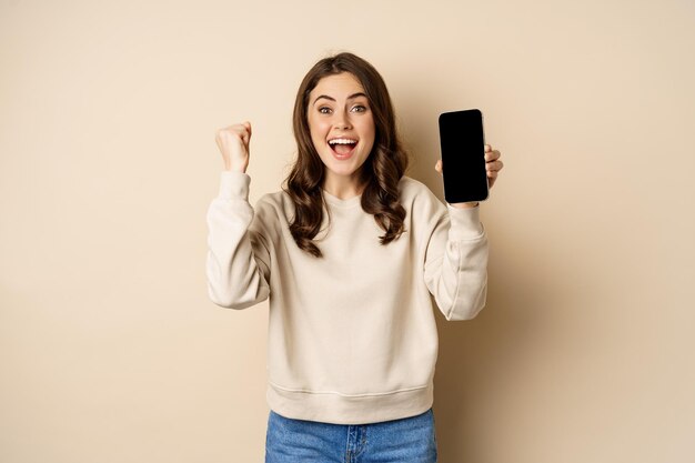 Enthusiastic woman cheering and showing cellphone screen smartphone app interface standing over beig...