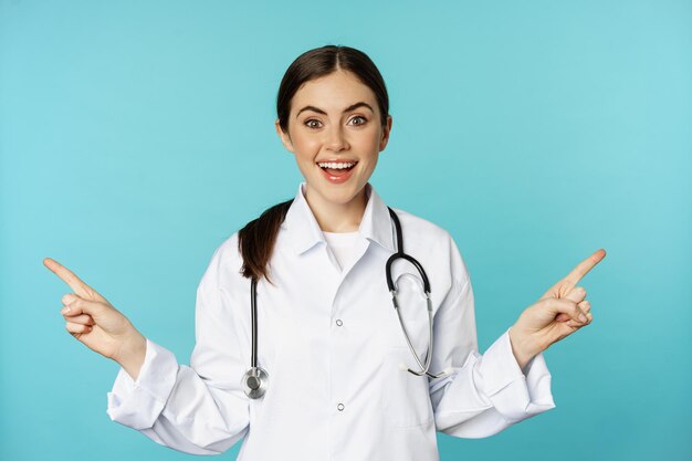 Enthusiastic woman doctor medical worker in white coat pointing fingers sidweays left and right show...