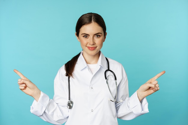 Восторженная женщина-врач, медицинский работник в белом халате, указывая пальцами в стороны, влево и вправо, показывая рекламу поликлиники, стоя на бирюзовом фоне