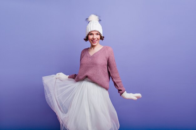 Enthusiastic woman in woolen mittens plays with her skirt isolated on purple wall. glad girl in hat and white gloves.
