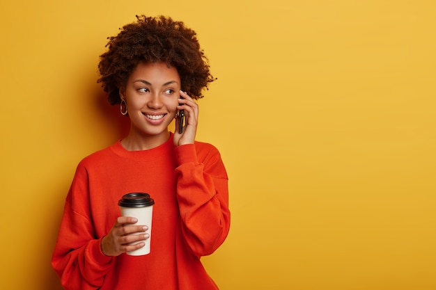 Entusiasta giovane ragazza afro ha una conversazione telefonica, parla con un amico e sente scherzi divertenti, vestita con abiti casual