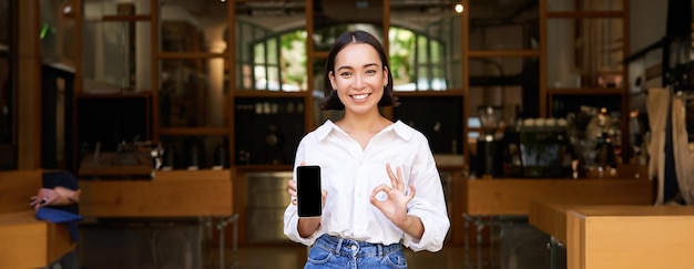 Foto gratuita appassionata giovane cameriera asiatica del caffè che mostra l'applicazione del telefono cellulare indicando