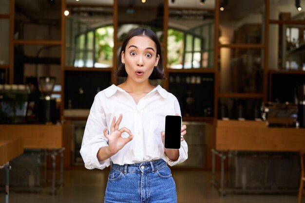 Free photo enthusiastic young asian girl waitress from cafe showing mobile phone application pointing at