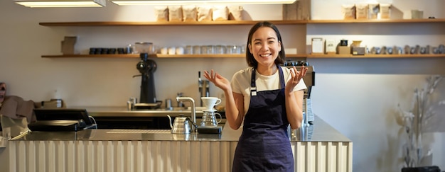 無料写真 エプロンを着た熱心なアジアの若い女性バリスタが驚いて手をたたき、笑顔で立っているのを見て