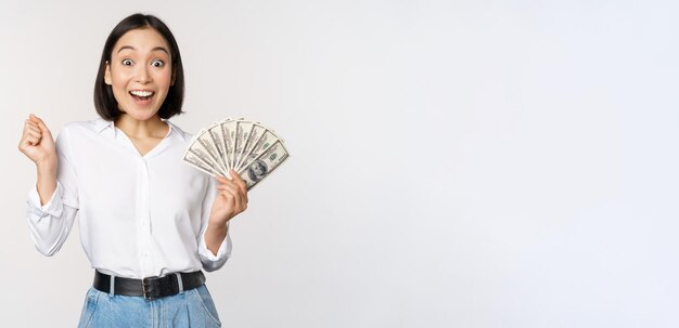 Восторженная молодая азиатка взволнованно смотрит в камеру, держа в руке деньги в долларах, стоя на белом фоне