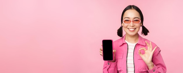 Free photo enthusiastic young asian woman showing okay ok sign smiling pleased mobile phone screen smartphone application standing over pink background