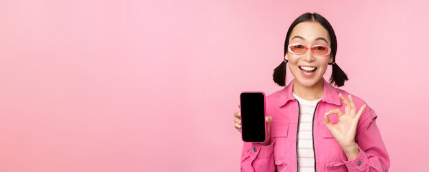 열성적인 젊은 아시아 여성이 분홍색 배경 위에 서 있는 만족스러운 휴대전화 화면 스마트폰 애플리케이션을 웃고 있는 OK 사인을 보여주고 있다