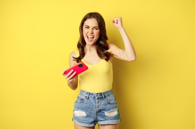 Enthusiastic young beautiful girl holding smartphone horizontally, rejoicing, winning on mobile phone video game, standing against yellow background.