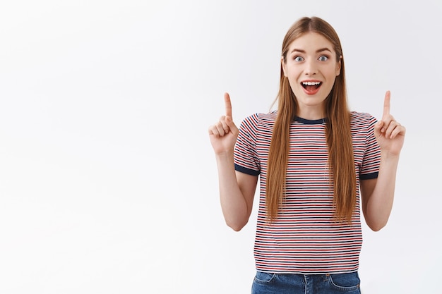 Enthusiastic young caucasian woman in striped t-shirt open mouth, gasping fascinated looks at camera intrigued and amused, pointing up, checking out amazing promo, standing on white wall stunned