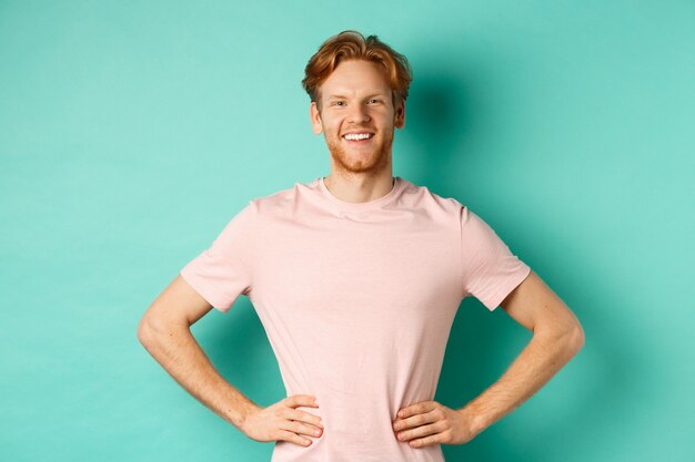 Enthusiastic young man with red hair, wearing t-shirt, standing happy and proud with hands on hops, standing over turquoise background.