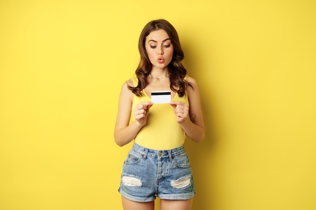 黄色の背景の上に立って、クレジットカードを示し、銀行を推薦し、店で割引、非接触型決済またはキャッシュバックを示す熱狂的な若い女性。