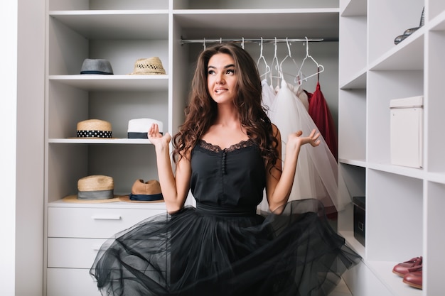 Бесплатное фото Восторженная молодая женщина, стоящая в раздевалке, шкафу и думающая, задумчиво смотрит. ее красивое черное платье парит в воздухе. у нее длинные вьющиеся каштановые волосы.