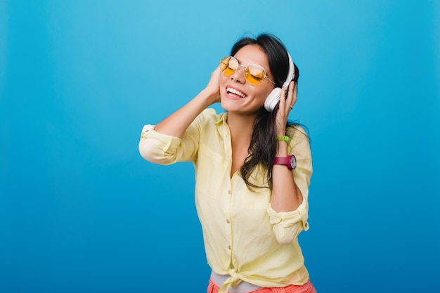 Восторженная молодая женщина в стильной желтой рубашке и розовом браслете трогательно наушниками, наслаждаясь песней. Фотография в помещении блаженной латиноамериканской девушки с блестящими темно-каштановыми волосами позирует.