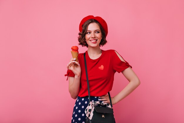 アイスクリームを食べる流行の服を着た熱狂的な若い女性。デザートでのんきな女性の笑顔の屋内写真。