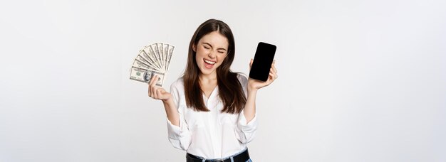 Free photo enthusiastic young woman winning money showing smartphone app interface and cash microcredit prize concept standing over white background