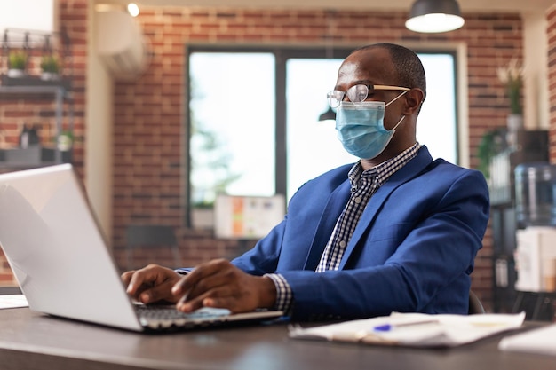 Entrepreneur looking at laptop screen to work on business project during pandemic. Employee wearing face mask and working on computer to plan marketing strategy in startup office.