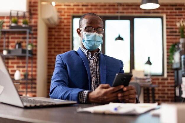 スマートフォンの画面を見て、営業所で仕事でフェイスマスクを着用している起業家。コロナウイルスのパンデミック時にデスクでタッチスクリーンを備えた携帯電話で作業している会社の従業員。