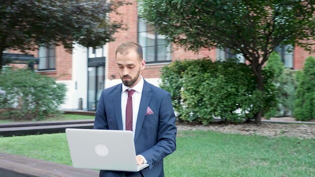 기업 전략을 입력하는 비즈니스 정보를 검색하는 노트북 컴퓨터를 들고 벤치에 앉아 있는 기업가 남자. 시작 건물 사무실 앞에 서 있는 사업가. 아웃도어 컨셉