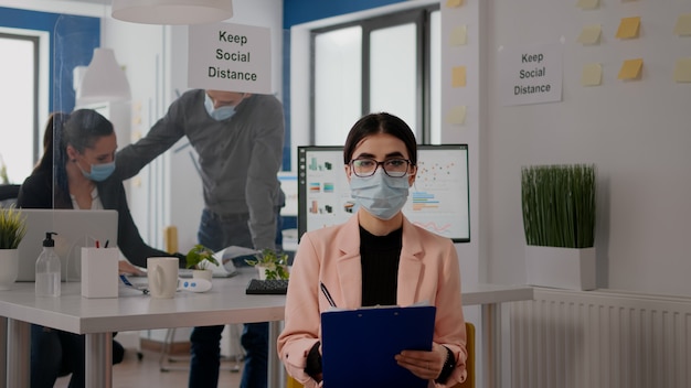 Entrepreneur sitting in new normal office looking into camera while writing lifestyle information during online videocall meeting. Businesswoman wearing face mask to avoid infection with covid19