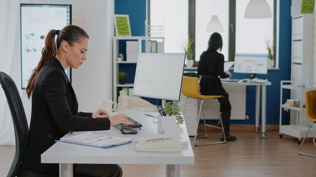 Entrepreneur using computer for business work with data charts on paper and monitor at corporate office. Businesswoman working with ideas for marketing strategy and management planning