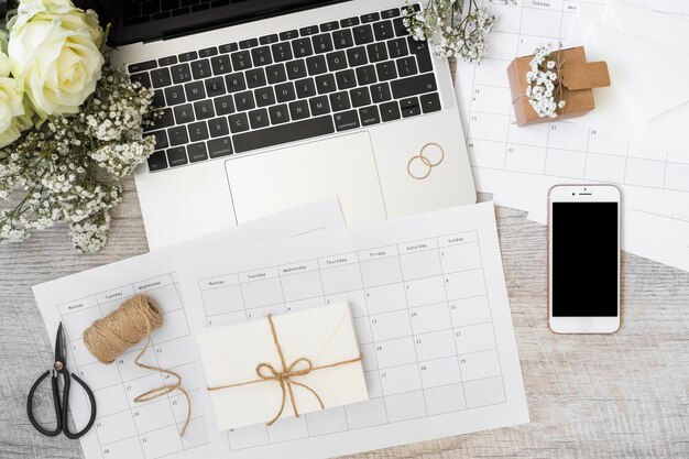 Envelope; calendar; laptop; flowers; smart phone; spool; scissor and spiral notebook on wooden desk