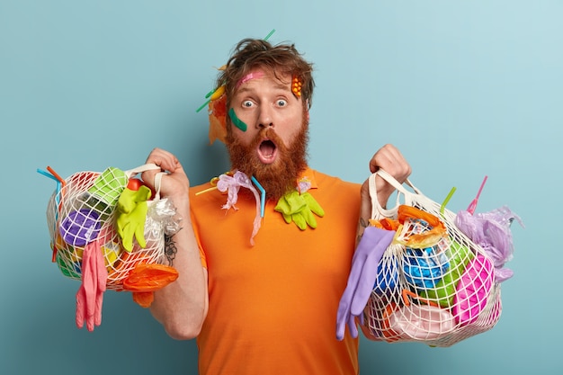 Environment protection concept. Puzzled shocked redhead bearded man stares, astonished by nature disaster, collects plastic rubbish, models over blue wall with two bags of garbage