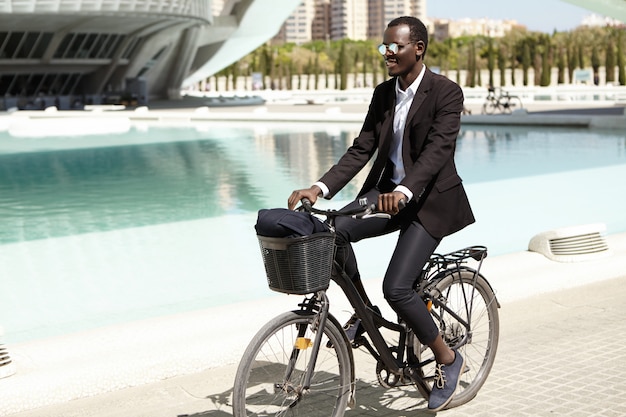 Environmentally friendly Afro American banker in formal wear and shades looking happy and relaxed, cycling to work on bike in urban setting, smiling cheerfully. Businesspeople, lifestyle and transport