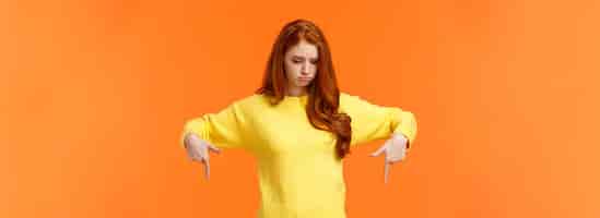 Foto gratuita invidia la donna rossa carina triste e imbronciata in maglione giallo che guarda e indica verso il basso con gelosia o d