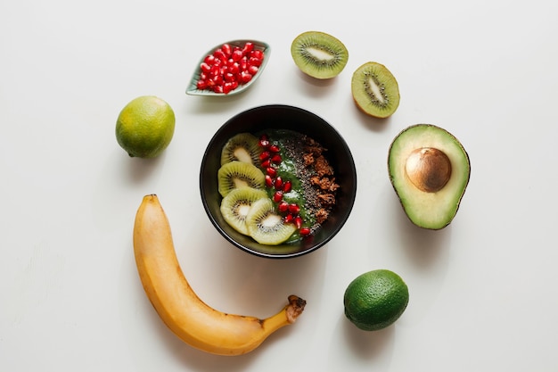 Essentials for cooking smoothie bowl. Blake plate topped with Kiwi, banana, pomegranate seeds, lime, granola, chia seeds. Healthy breakfast.  Round composition.