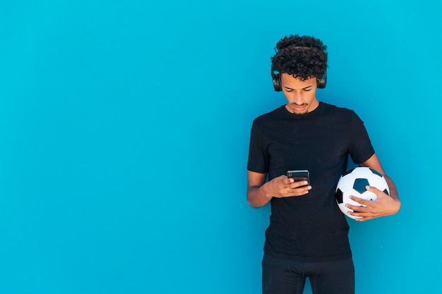 Бесплатное фото Этнические кудрявый спортсмен, держа футбол и с помощью телефона