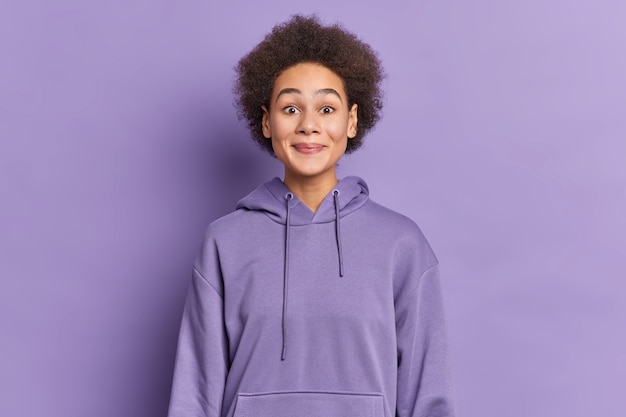 ethnic girl with Afro hair smiles pleasantly and looks curiously wears hoodie.