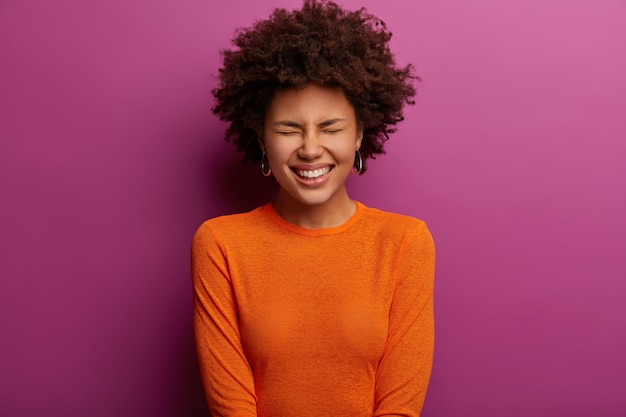 Ethnic overjoyed young woman laughs at something positive, closes eyes and giggles happily, dressed in orange jumper, hears funny joke, poses against vibrant purple wall. Emotions concept