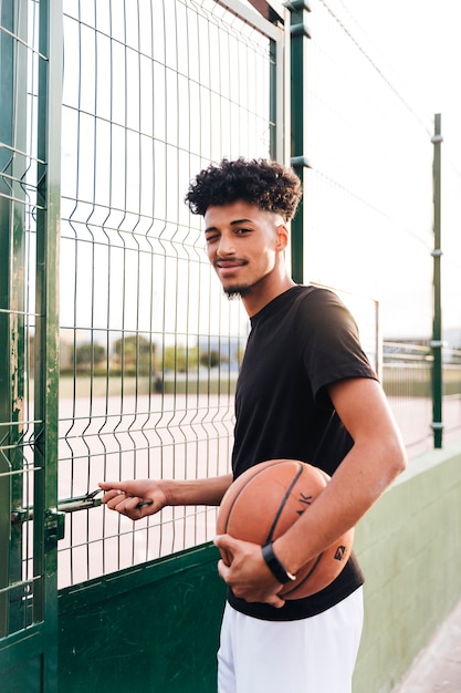 Этнические молодой человек, подмигивая, стоя с баскетболом