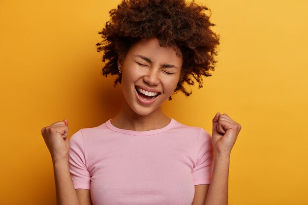 Euphoric curly woman makes yes gesture, clenches fists, excited by great news, celebrate victory, has overjoyed expression, got prize, tilts head, poses and gestures against yellow wall