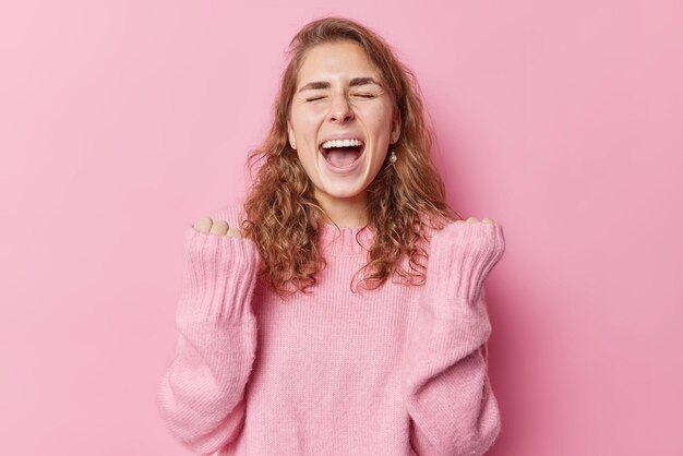 Euphoric overjoyed long haired woman keeps mouth widely opened exclaims from joy clenches fists with triumph celebrates achhievement wears warm sweater isolated over pink background reaches goal