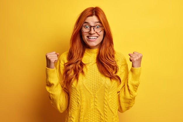 Euphoric overjoyed redhead woman raises clenched fists makes yes gesture excited by excellent news wears spectacles and yellow jumper celebrates getting prize stands indoor. Success victory concept