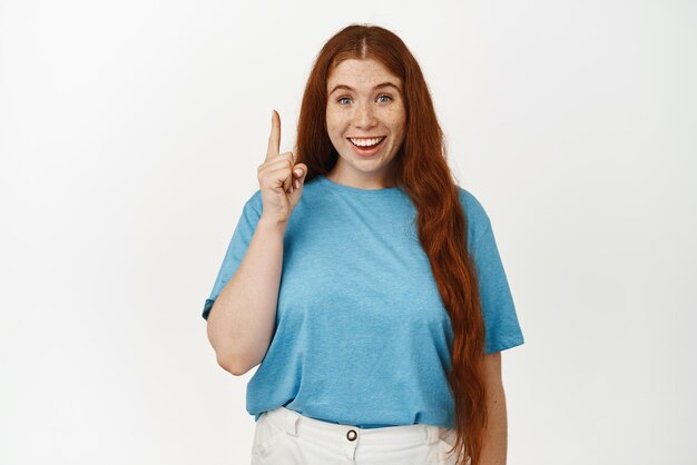 Eureka Excited redhead girl with candid smile pointing finger up pitching an idea got something interesting showing advertisement on top standing happy against white background