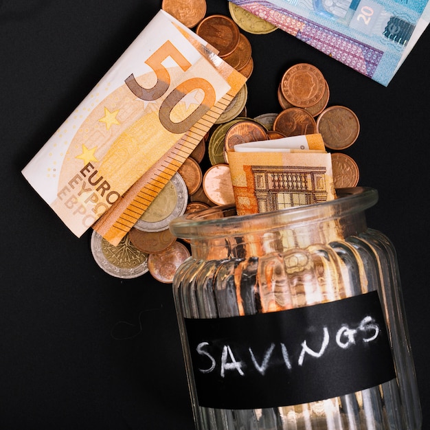 Евро банкноты и монеты, пролитые из открытой банки сберегательного стекла на черном фоне