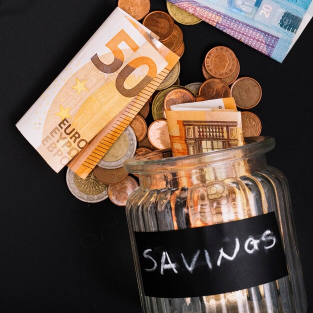 Euro banknotes and coins spilling from open savings glass jar on black background